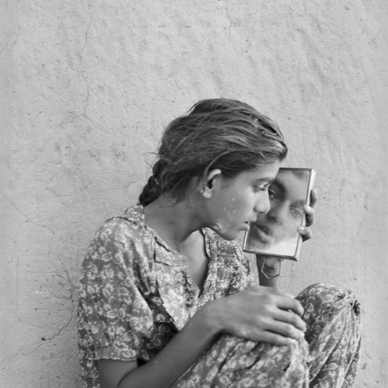 B/W image of a sitting young woman looking at herself in a mirror she holds in her left hand